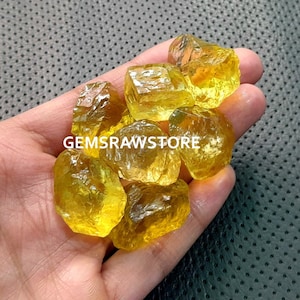 May include: A collection of yellow citrine crystals, with rough, faceted surfaces. The crystals are translucent and have a slight golden hue. The crystals are arranged in a hand, with the text "GEMSRawSTORE" visible in the image.