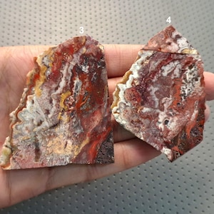 Puede incluir: Dos losas de piedra pulida de forma irregular. Las piedras muestran una mezcla de colores, incluyendo rojo, blanco, amarillo y marrón, con patrones y texturas intrincados. Las losas se sostienen en una mano, mostrando su belleza natural.
