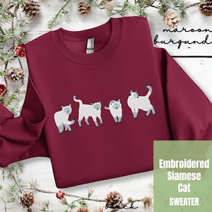 May include: A maroon sweatshirt with an embroidered design of four Siamese cats. The text "Embroidered Siamese Cat SWEATER" is printed on the bottom right corner of the sweatshirt.