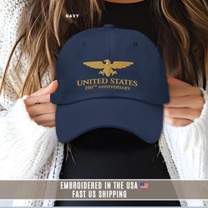Navy blue embroidered United States 250th anniversary baseball cap with gold eagle design, held by model, front view.
