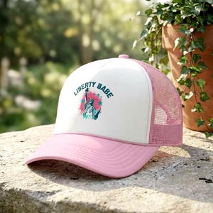 May include: A pink and white trucker hat with the words "LIBERTY BABE" printed on the front. The design features an illustration of the Statue of Liberty. The hat has a pink brim and mesh back.