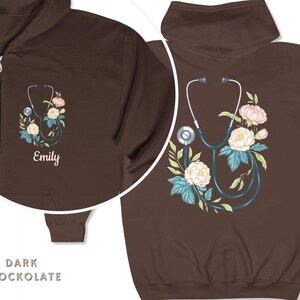 Pu&ograve; includere: Felpa con cappuccio con zip di colore cioccolato scuro con stetoscopio e design floreale. Il testo "Emily" &egrave; ricamato sulla parte anteriore della felpa.
