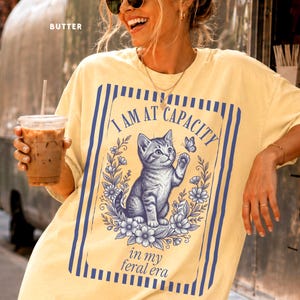 Funny Cat Shirt on a Butter Comfort Colors 1717 tee featuring an I Am at Capacity kitten graphic with in my feral era text, styled in a New York coffee van lifestyle mockup for cat lovers, introverts, overstimulated friends, and funny gift shoppers.