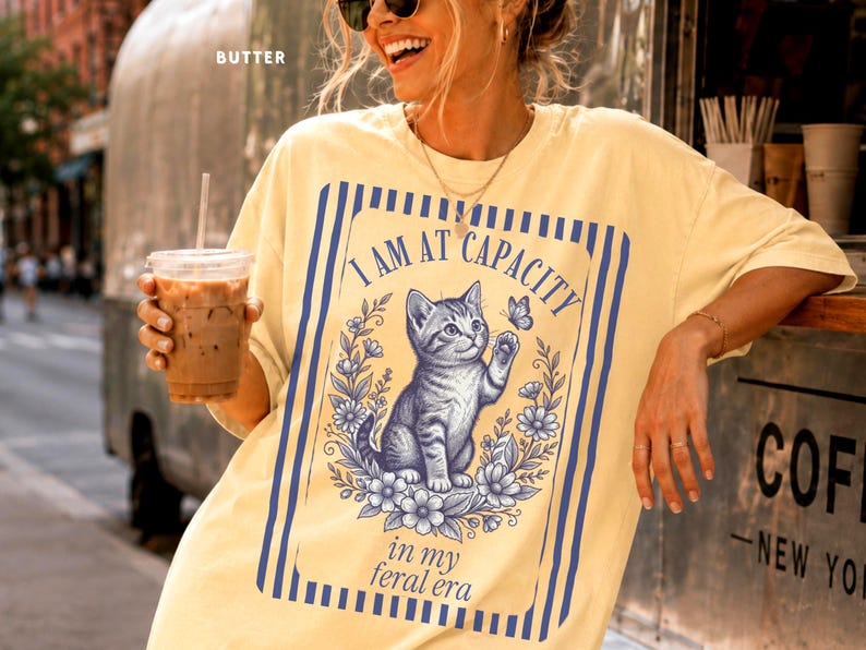 Funny Cat Shirt on a Butter Comfort Colors 1717 tee featuring an I Am at Capacity kitten graphic with in my feral era text, styled in a New York coffee van lifestyle mockup for cat lovers, introverts, overstimulated friends, and funny gift shoppers.