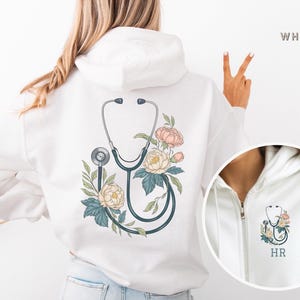 Pu&ograve; includere: Felpa con cappuccio bianca con un design floreale e uno stetoscopio sul retro. La felpa ha una cerniera e un cappuccio. Il testo "HR" &egrave; sul davanti della felpa.
