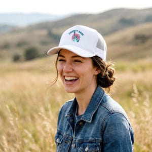May include: White baseball cap with the text "LIBERTY BABE" in blue and pink, with a floral design. The cap has a mesh back and a curved brim. The person in the image is wearing a denim jacket.