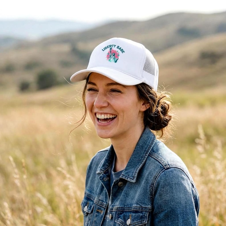 May include: White baseball cap with the text "LIBERTY BABE" in blue and pink, with a floral design. The cap has a mesh back and a curved brim. The person in the image is wearing a denim jacket.