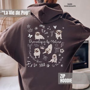 May include: Dark chocolate zip hoodie featuring a pug-themed design. The back of the hoodie displays pug illustrations, the phrase "Dramatique by Nature," and the text "La Vie de Pug." Additional text includes "Certified Diva Queen" and "XOXO."