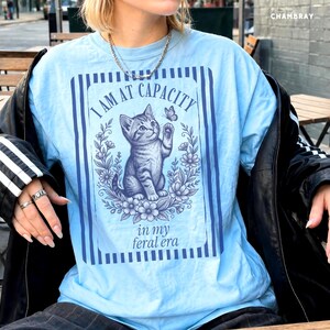 I Am at Capacity shirt on a Chambray Comfort Colors 1717 tee with a funny kitten graphic and feral era quote, shown in a coffee shop bistro lifestyle mockup for cat moms, introverts, coffee girls, and chaotic best friends.