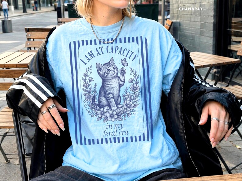 I Am at Capacity shirt on a Chambray Comfort Colors 1717 tee with a funny kitten graphic and feral era quote, shown in a coffee shop bistro lifestyle mockup for cat moms, introverts, coffee girls, and chaotic best friends.
