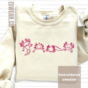 May include: A cream-coloured sweatshirt with a crew neck and long sleeves. The front displays an embroidered design of four pink axolotls in different poses. The text "Embroidered sweater" is in a white box in the lower right corner.