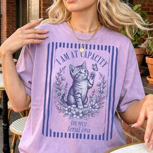 Overstimulated Shirt on an Orchid Comfort Colors tee featuring a funny cat graphic with I Am at Capacity and in my feral era text, styled as a relaxed oversized tee for chaotic girls, introverts, coffee lovers, and cat mom gifts.