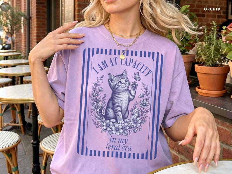 Overstimulated Shirt on an Orchid Comfort Colors tee featuring a funny cat graphic with I Am at Capacity and in my feral era text, styled as a relaxed oversized tee for chaotic girls, introverts, coffee lovers, and cat mom gifts.