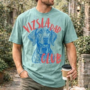 May include: A sage green short-sleeve t-shirt featuring a blue line-art illustration of a Vizsla dog face in the center. The text "VIZSLA DAD CLUB" is printed in a bold, red, hand-lettered style font arched over and below the dog graphic.