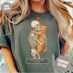 May include: Dark green t-shirt with a skeleton hugging an orange tabby cat. The graphic includes the text "Purr-eternity No.19". The shirt has a vintage, faded appearance and is labelled "Comfort Colors" and "More Colors".