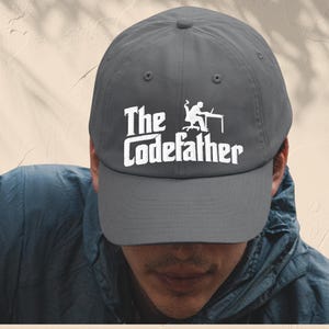 May include: A grey baseball cap with the white text "The Codefather" and a graphic of a person at a computer. The cap is worn by a person in a blue jacket.