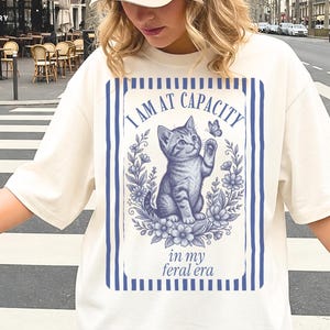 Funny Cat Lover Shirt on an Ivory Comfort Colors 1717 tee with an I Am at Capacity and in my feral era kitten design, shown in a casual streetwear mockup for introverts, cat moms, college students, and funny friend gifts.