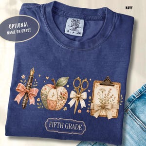 May include: A navy blue t-shirt featuring a graphic print of a fountain pen with a pink bow, a patchwork apple, gold scissors with a white bow, and a clipboard with floral art. The text reads FIFTH GRADE and OPTIONAL NAME OR GRADE.