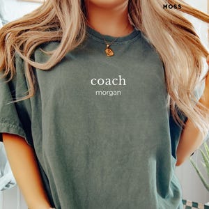 Puede incluir: Una camiseta verde salvia con la palabra "coach" y el nombre "morgan" impresos en blanco. La camiseta tiene mangas cortas y un corte holgado. La palabra "MOSS" está impresa en la esquina superior derecha.