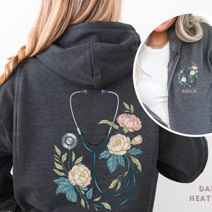 Pu&ograve; includere: Felpa con cappuccio color grigio scuro melange con un design floreale e uno stetoscopio sul retro. Il testo "Amelia" &egrave; ricamato sul davanti della felpa.