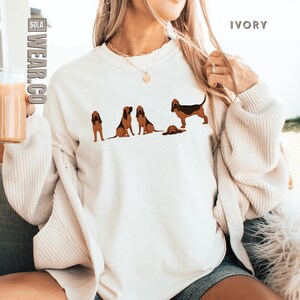 May include: A white sweatshirt with a graphic of five brown and black Bloodhounds. The sweatshirt is on a person wearing a cream cardigan and denim shorts. The text "SOLA WEAR.CO" is in the top left corner of the image and the text "IVORY" is in the top right corner of the image.