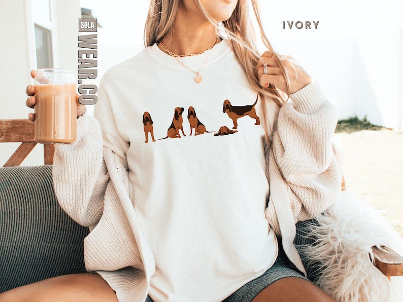 May include: A white sweatshirt with a graphic of five brown and black Bloodhounds. The sweatshirt is on a person wearing a cream cardigan and denim shorts. The text "SOLA WEAR.CO" is in the top left corner of the image and the text "IVORY" is in the top right corner of the image.