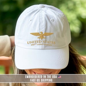 White embroidered United States 250th anniversary cap with gold eagle design, worn outdoors showing front stitching.
