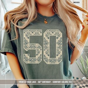 Peut inclure: Un t-shirt à manches courtes vert mousse arborant un grand graphique crème représentant le nombre 50. Le nombre 50 est rempli d'un motif de fausse dentelle détaillé et complexe. Le texte imprimé en bas indique : PRINTED FAUX LACE - 50TH BIRTHDAY - COMFORT COLORS TEE.
