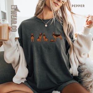 May include: A woman wearing a dark gray t-shirt with a graphic of four bloodhounds. The shirt has a distressed look and the graphic is in shades of brown and black.