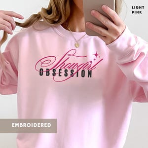 Puede incluir: Sudadera rosa claro bordada con la frase "Showgirl Obsession" en cursiva y una estrella. El texto "Light Pink" está en la esquina superior derecha. La palabra "Embroidered" está en la parte inferior.
