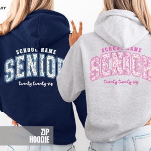 May include: A navy blue hoodie and an ash gray hoodie, both featuring a back graphic with the text "SCHOOL NAME" above the word "SENIOR" in a floral pattern, and "twenty twenty six" in script below. The navy hoodie has blue floral text, while the ash gray hoodie has pink floral text.
