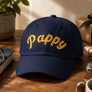 May include: A navy blue baseball cap featuring the word Pappy embroidered in yellow script lettering across the front panel. The cap has a structured crown, a curved brim, and a button at the top center, showcasing a classic six-panel design.