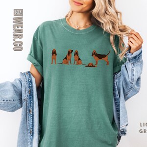 May include: A light green t-shirt with a graphic of five Bloodhound dogs. The dogs are in various poses, including sitting, standing, and running. The text "SOLA WEAR.CO" is printed on the left side of the shirt.