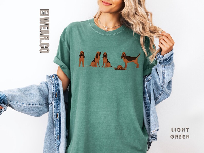 May include: A light green t-shirt with a graphic of five Bloodhound dogs. The dogs are in various poses, including sitting, standing, and running. The text "SOLA WEAR.CO" is printed on the left side of the shirt.