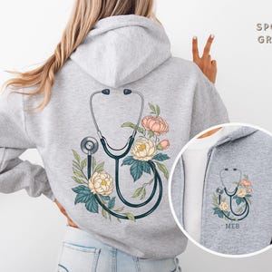 Pu&ograve; includere: Una felpa con cappuccio grigia con un design floreale e uno stetoscopio sul retro. La felpa &egrave; mostrata su un manichino. Il testo "Sport Grey" &egrave; visibile nell'angolo in alto a destra dell'immagine.
