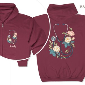 Pu&ograve; includere: Felpa con cappuccio con zip color bordeaux con uno stetoscopio e un design floreale. Il testo "Emily" &egrave; ricamato sulla parte anteriore della felpa.