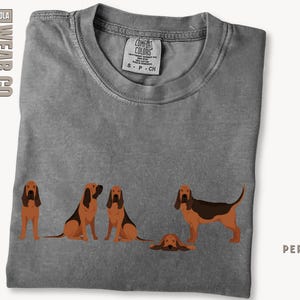 May include: A gray t-shirt with a graphic of four Bloodhound dogs. The dogs are in various poses, including sitting, standing, and lying down. The text "COMFORT COLORS" is printed on the tag.