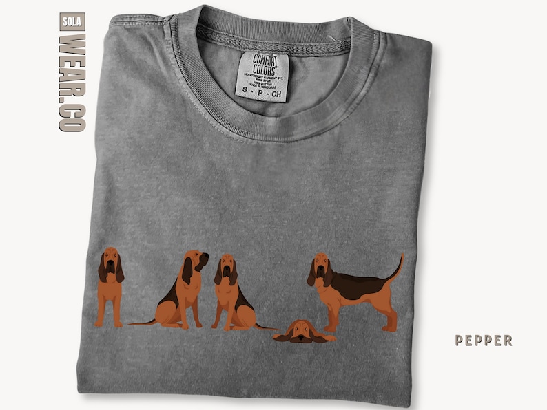 May include: A gray t-shirt with a graphic of four Bloodhound dogs. The dogs are in various poses, including sitting, standing, and lying down. The text "COMFORT COLORS" is printed on the tag.