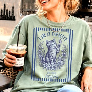Cat Mom Shirt on a Bay Comfort Colors 1717 tee with a funny I Am at Capacity kitten graphic and feral era quote, shown in a New York coffee van lifestyle mockup for cat lovers, social battery humor, introvert gifts, and casual outfits.