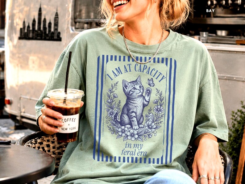 Cat Mom Shirt on a Bay Comfort Colors 1717 tee with a funny I Am at Capacity kitten graphic and feral era quote, shown in a New York coffee van lifestyle mockup for cat lovers, social battery humor, introvert gifts, and casual outfits.