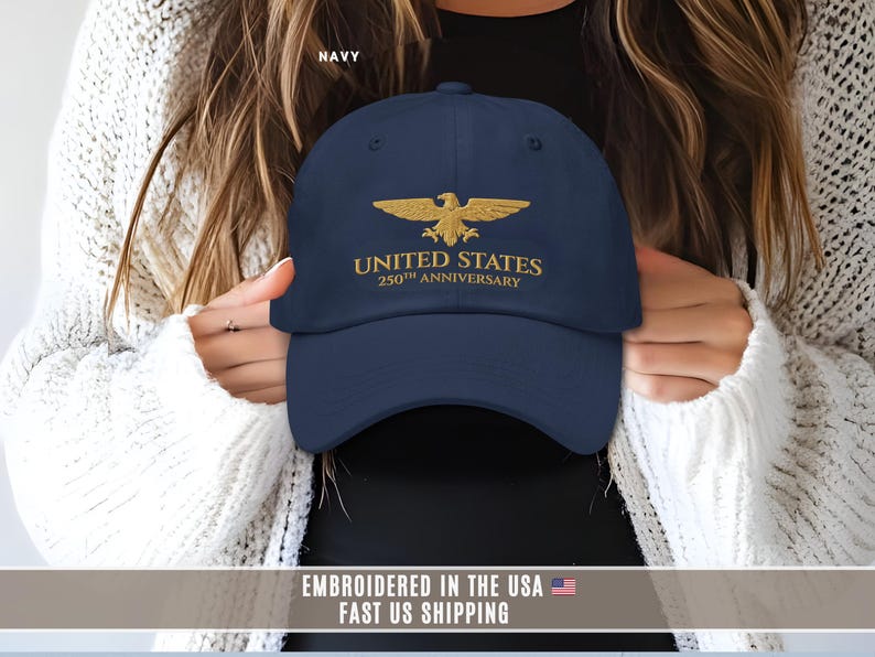 Navy blue embroidered United States 250th anniversary baseball cap with gold eagle design, held by model, front view.