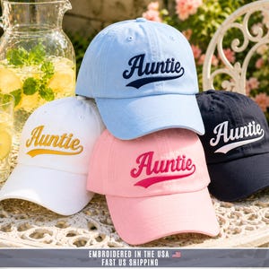 May include: A collection of four baseball caps in light blue, white, pink, and navy blue. Each cap features the word Auntie embroidered in a cursive script font on the front panel. The light blue cap has navy text, the white cap has gold text, the pink cap has dark pink text, and the navy cap has white text.