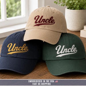 May include: Three baseball caps in tan, navy blue, and dark green. Each cap features the word Uncle embroidered in a cursive script font on the front panel. The tan cap has maroon embroidery, the navy cap has gold, and the green cap has white.