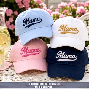 May include: Four baseball caps in light blue, white, pink, and navy blue. Each cap features the word Mama embroidered in a cursive script font on the front panel. The light blue cap has dark blue text, the white cap has gold text, the pink cap has dark pink text, and the navy blue cap has white text.