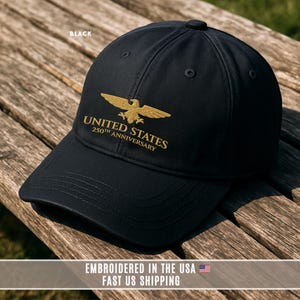 Black embroidered United States 250th anniversary baseball cap with gold eagle logo displayed on rustic wooden bench.