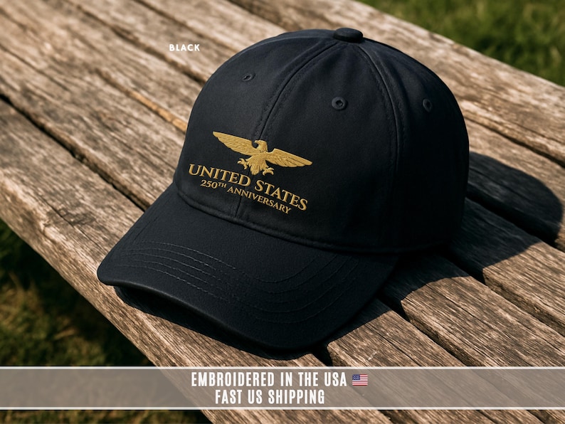 Black embroidered United States 250th anniversary baseball cap with gold eagle logo displayed on rustic wooden bench.