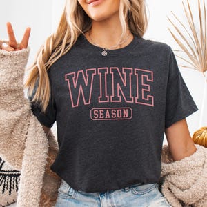 Peut inclure: T-shirt gris fonc&eacute; avec l'inscription "WINE SEASON" en lettres roses. Le t-shirt est de style col rond. Le mod&egrave;le porte un short en jean et un cardigan beige.