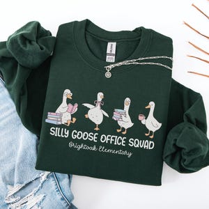 May include: A dark green sweatshirt with the words "SILLY GOOSE OFFICE SQUAD" and "Brightoak Elementary" printed on the front. The graphic features four cartoon geese. A silver necklace and blue jeans are also in the image.
