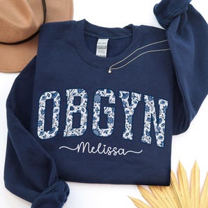 May include: A navy blue sweatshirt with the letters OBGYN in a floral pattern. The name "Melissa" is written in cursive below the letters. A gold necklace with a star pendant is visible.