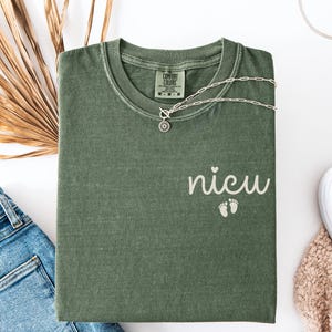 May include: A moss green t-shirt with the word "nicu" and two baby footprints printed on the front. A silver necklace with a pendant is draped over the shirt. The word "Moss" is printed at the bottom of the image.